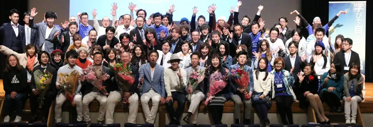 1000 人セミナーの会場で、壇上に並んだ参加者たちが手を挙げている集合写真